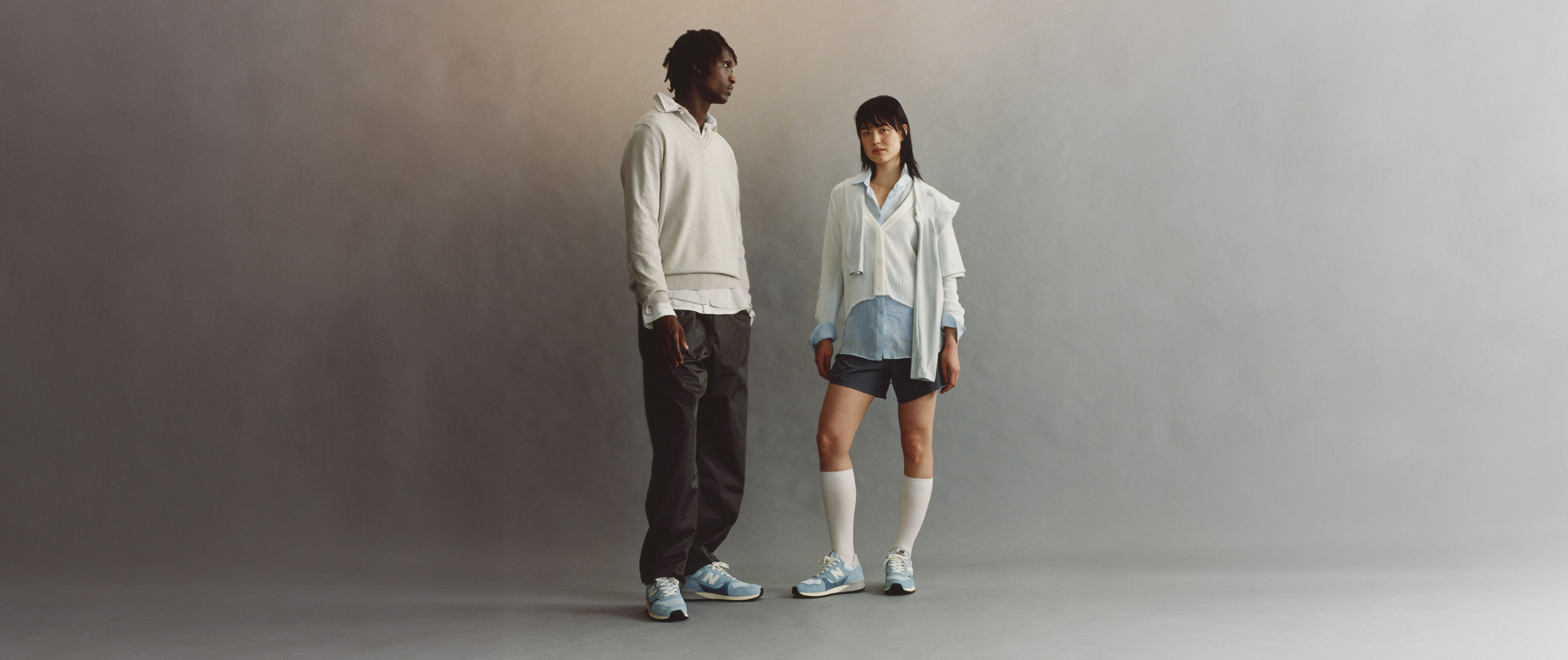 A man and a woman standing on a grey studio background, both wearing New Balance 475 shoes.