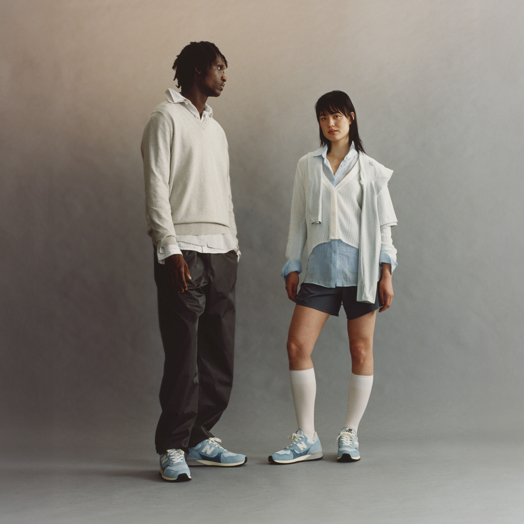 A man and a woman standing on a grey studio background, both wearing New Balance 475 shoes.